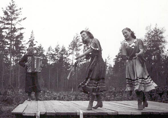 Metsn rakennettu esiintymislava, jossa tanssi kaksi liehuviin pukuihin sonnustautunutta naista. Taustalla sotilas soittaa hanuria.